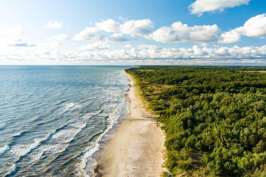 Aerial view of the coast of the Baltic Sea. To the right are dense green forests, in the center a white sandy beach runs through the picture, and on the left is the sea. Waves break along the coast. The sky is blue with some clouds.