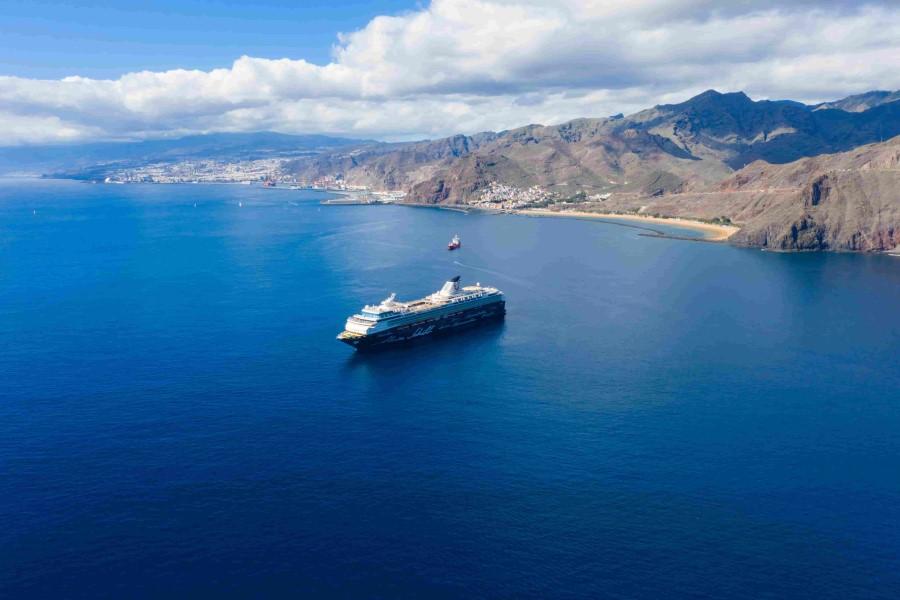 A cruise ship sails in sunny weather on the blue sea along a mountainous coast that popular cruise routes pass. In the background, a city and another ship lie anchored.
