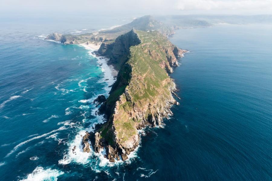 Aerial view of a rugged coastline with green hills extending into the sea. The cliffs are surrounded by turquoise, choppy water. Perfect scenery for the popular cruise routes.