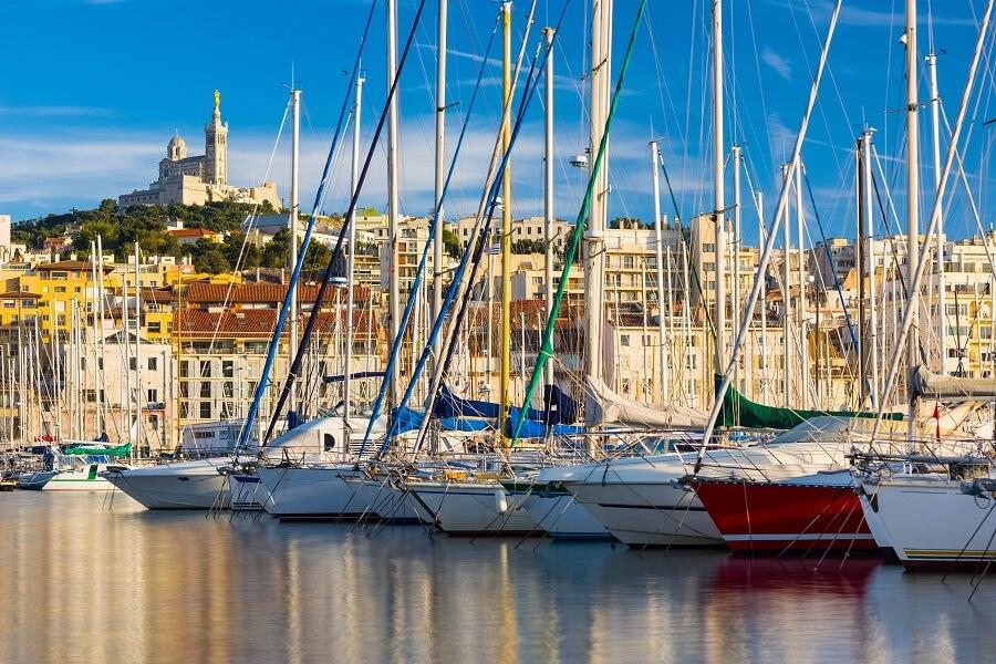 Sailing boats in the harbor of Marseille, France, with the Basilica Notre-Dame de la Garde on a hill in the background. Blue and green mastboats dominate the foreground while the city's buildings line the center.