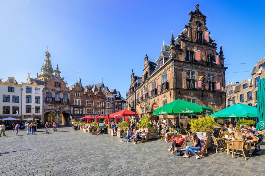 Paved square in Nijmegen with a historical building on the right, lively cafés, and colorful umbrellas in red and green on the left. People sit relaxed in sunny weather.