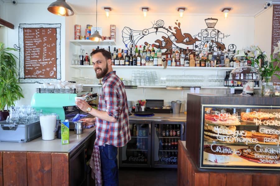 A man in a checkered shirt stands at the counter of a cozy café, surrounded by glasses and bottles. In the background, an artistic mural featuring coffee cups and inviting lighting.